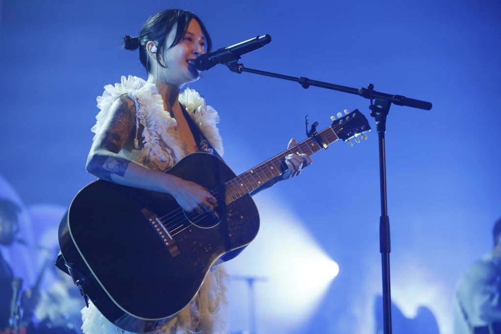 Japanese Breakfast's Michelle Zauner smiles while playing a black acoustic guitar and singing, looking out over a sold-out crowd at The Sylvee. She's wearing a white dress that is nearly all frills. Behind her, the stage is cast in a cool blue light.