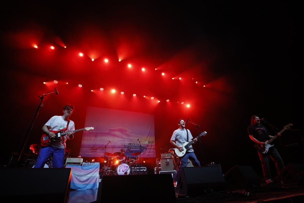 A wide shot of the four members of PUP performing at The Sylvee. A trans flag is proudly displayed over a guitar amp on the center left, a banner displaying album art covers a significant portion of the black wall, and all of the members' faces are contorted as they play and sing. They are bathed in blood red light.