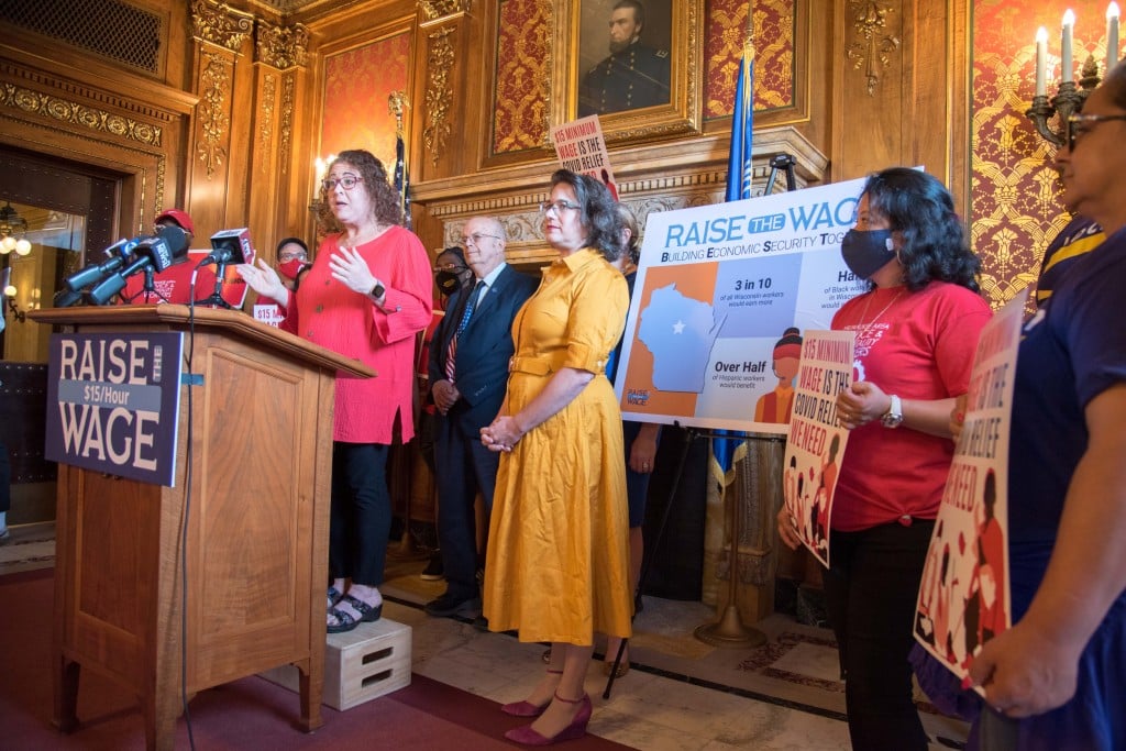 Angled photo of a press conference from mid-2021 shows several women and men standing in an ornately decorated government building. A woman in a salmon-colored shirt stands at a lectern and speaks into several microphones that sit on top. Signs displaying "Raise The Wage $15/Hour" and "$15 Minimum Wage Is The Covid Relief We Need" are displayed and held up around her.