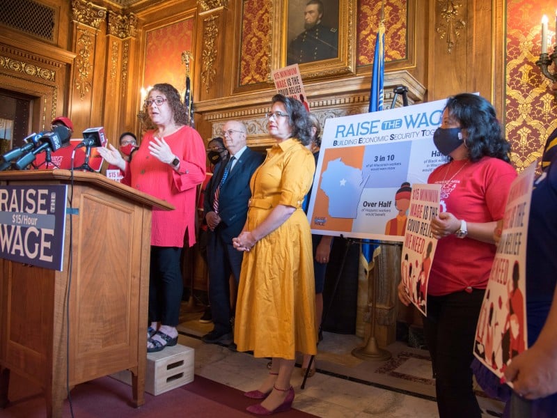 Angled photo of a press conference from mid-2021 shows several women and men standing in an ornately decorated government building. A woman in a salmon-colored shirt stands at a lectern and speaks into several microphones that sit on top. Signs displaying "Raise The Wage $15/Hour" and "$15 Minimum Wage Is The Covid Relief We Need" are displayed and held up around her.