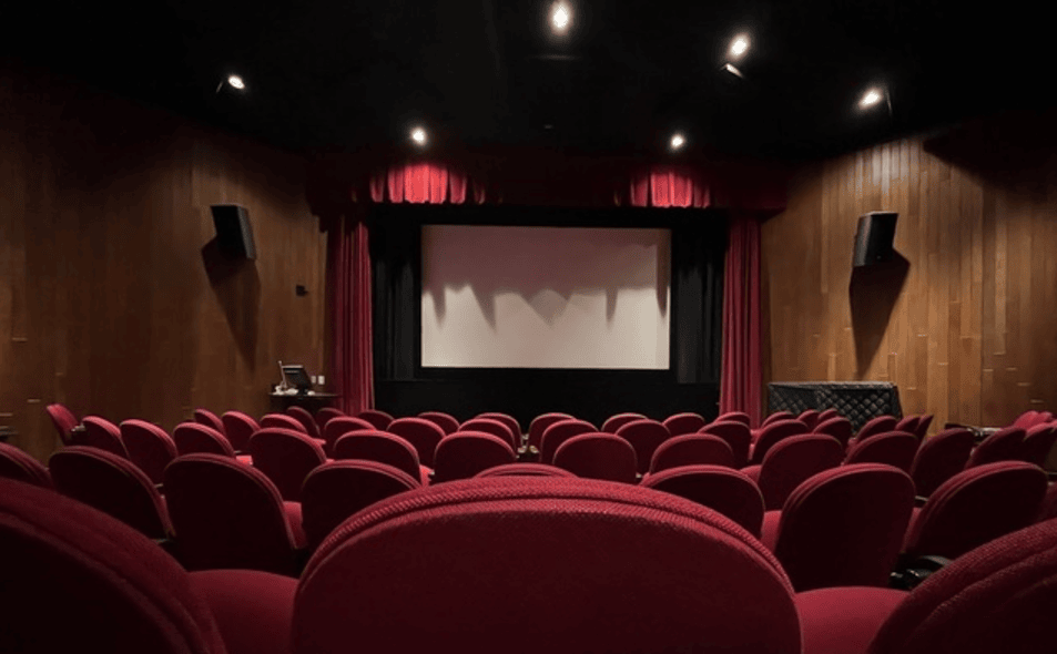 A wide-angled photo taken near the back of an empty movie theater. The red seats fan out in a U-shaped rows. The projection screen is shown with fluorescent lights casing shadows on it at the front of the room.
