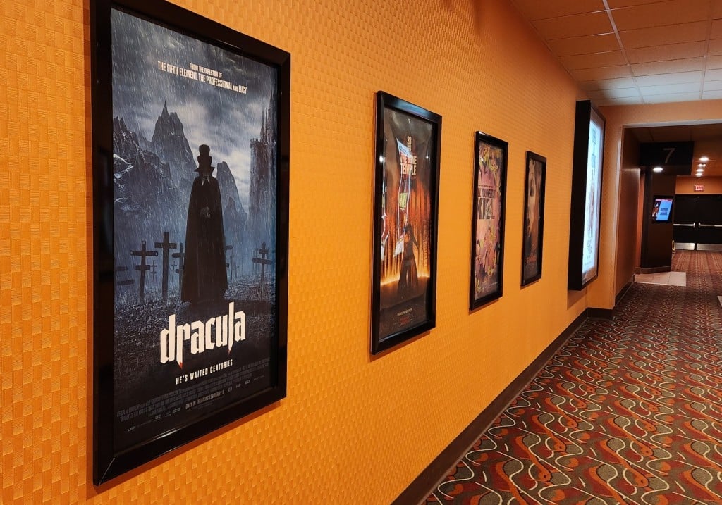 An angled photo in a movie theater hallway displays a row of framed posters hanging on the orange wall. The posters include "Dracula," "28 Years Later: The Bone Temple," and "All You Need Is Kill." A squiggly-patterned carpet can be seen below near the right edge of the photo.