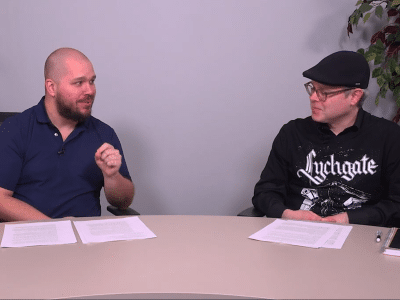Medium close-up video still of two people sitting at a table in a conference room. They are turned towards one another and engaged in conversation. The person on the left wears a dark blue polo shirt and makes a gesture with his left hand while he speaks. The person on the right smiles slightly, wearing a black Lychgate band shirt and black Kangol cap. He listens with his eyes angled down.