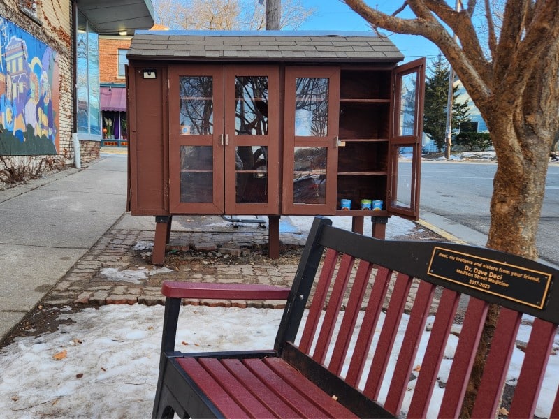 Daytime photo of the outdoor food pantry and bench on Few Street around the corner from the Social Justice Center in Madison. The wooden bench sits in the foreground at an angle with a small plaque on the back that reads "Rest, my brothers and sisters, from your friend, Dr. Dave Deci Madison Street Medicine 2017-2023." Behind it is a larger wooden cabinet with two sets of doors and glass panels that contains only a few canned goods on the shelving inside. The rightmost door is open.