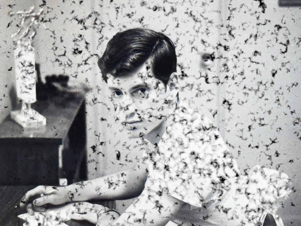 An old black-and-white photograph from the mid-late 1960s of a young man sitting at a desk in a room. He turns his gaze towards the camera and stares with a neutral expression. The photo is altered with black digital debris that litters the image and emulates mental noise.