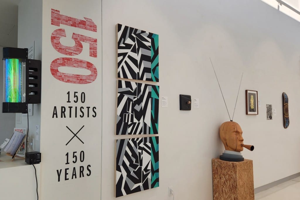 A slightly angled photo of an art gallery space with white walls. Various artworks large and small are displayed on the wall (and one a wooden pedestal on the marble floor). At the left, the number 150 is displayed vertically in a large pinkish-red font, while the exhibition name "150 ARTISTS X 150 YEARS" is displayed horizontally in smaller black font.
