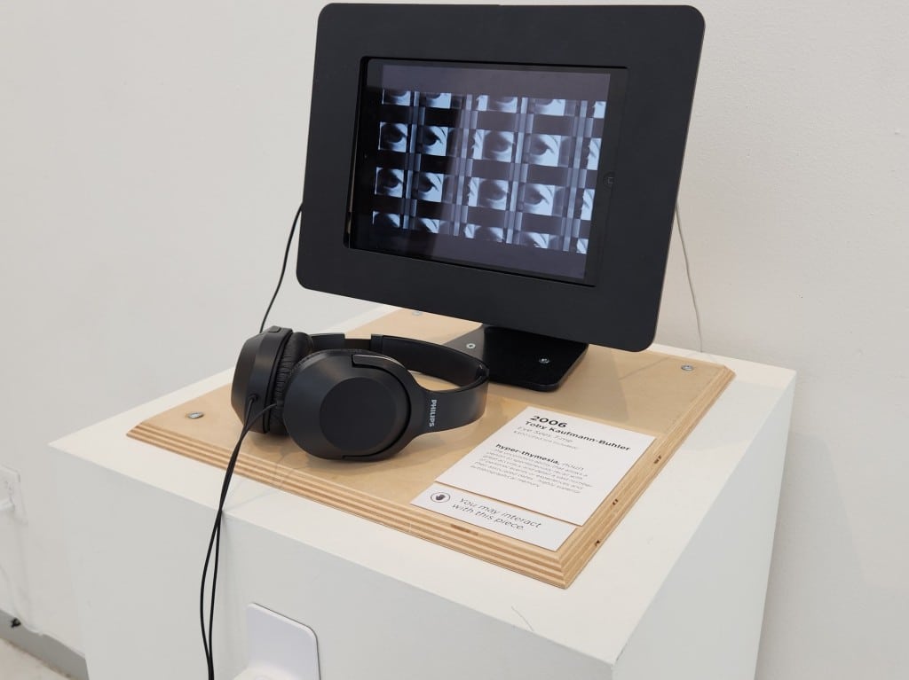 Angled photo of an iPad that is mounted on a larger black plastic display and screwed down to a wooden slate and white pedestal that displays the artist name "Toby Kaufmann-Buhler," the year of "2006" and the word prompt for the project, "hyper-thymesia." The iPad screen shows a video of horizontal rows of human eyes blinking at an extreme close-up. A pair of black headphones rests atop the pedestal.