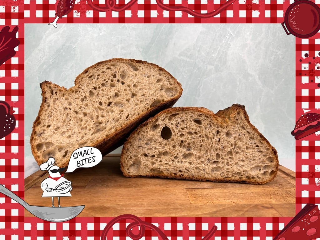 Photo of a freshly baked sourdough bread loaf, cut in half, sitting on a cutting board. The wall in the background is a pale-green marble color. Bordering this photo is a red and white checkered tablecloth illustration. In the lower left corner of this illustrated frame is a small chef with an oversized mustache standing on a spoon. The chef's speech bubble reads "Small Bites."