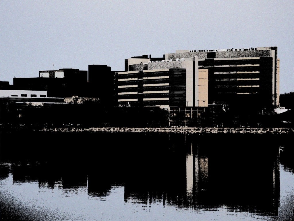 A color-altered photo taken from the westernmost edge of the University of Wisconsin–Madison campus and University Bay. UW Health University Hospital, the UW–Madison Health Sciences Learning Center, and the Wisconsin Institutes for Medical Research buildings cast heavy shadows onto Lake Mendota in the foreground.