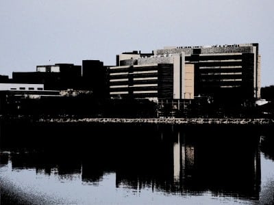 A color-altered photo taken from the westernmost edge of the University of Wisconsin–Madison campus and University Bay. UW Health University Hospital, the UW–Madison Health Sciences Learning Center, and the Wisconsin Institutes for Medical Research buildings cast heavy shadows onto Lake Mendota in the foreground.