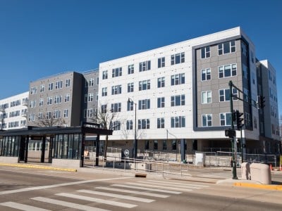 Photo of a large, boxy, multi-family housing complex at a street corner. It's painted in shades of white and grey. Each unit has thin, elongated sets of windows. The traffic intersection at a BRT (Bus Rapid Transit) stop can be seen in the foreground.