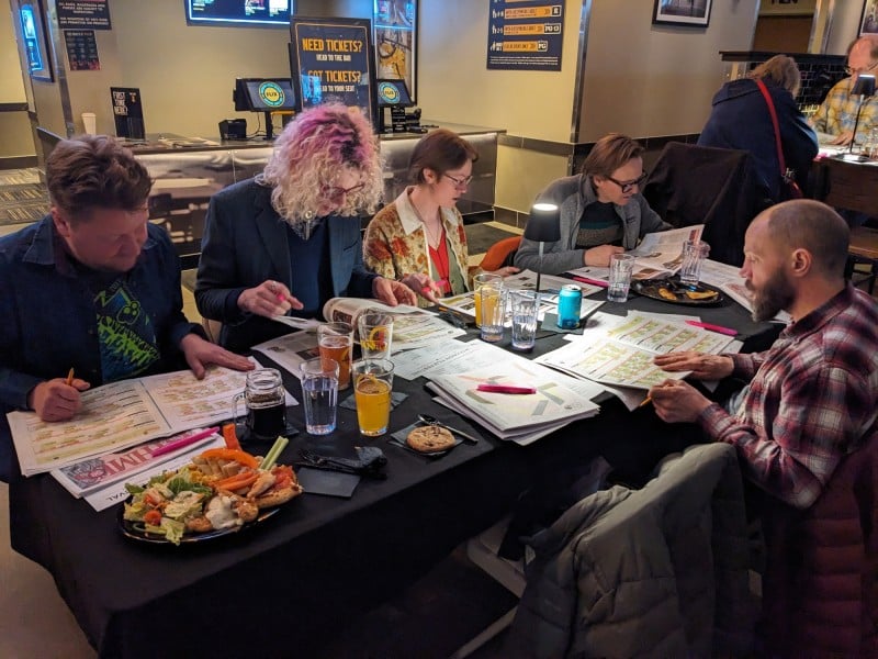 Angled photo at a medium shot of five people seated at a table in a theater lobby. Four people sit on the far side, while one person sits closest to the camera on the other side. They are all absorbed in the news-printed 2026 Wisconsin Film Festival guides on the table. The table contains numerous beverage glasses, a couple plates of food, and a small table lamp towards the center.