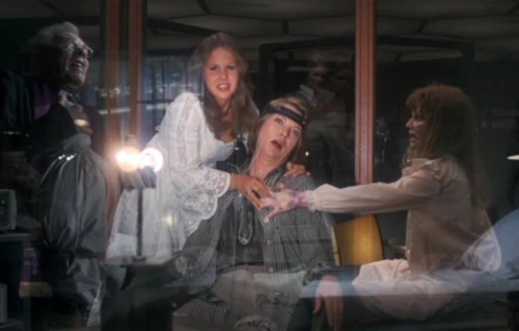 A still from the film "Exorcist II: The Heretic" captures an absurdist scene with various demonic forces, only partly transparent, haunting a middle-aged woman who is hooked up to a brain wave-monitoring machine. She stares off into the middle distance with her mouth agape.