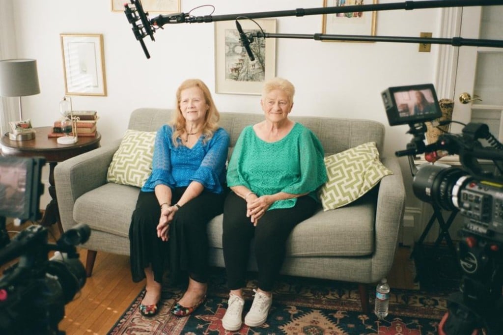 A production photo from Ken Kwapis' documentary "We Are The Shaggs" shows two elderly women sitting on a couch facing two visible film cameras in the foreground at the left and right corners of the frame. Both women smile in the sunlit room.