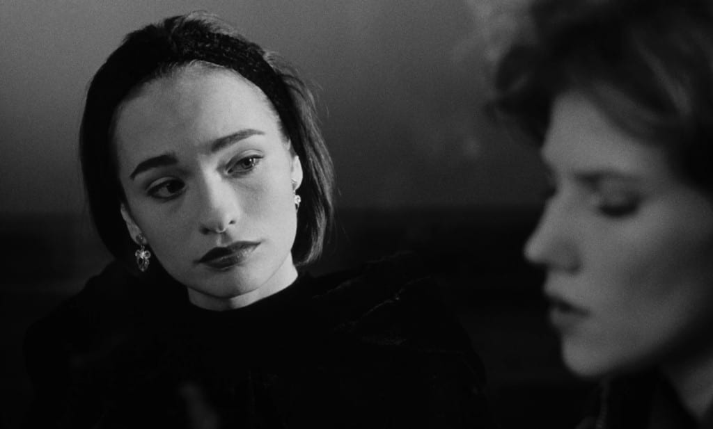 A black-and-white still from the film "Nadja" shows a young brunette actress at a medium shot. She looks towards the right of the frame where another woman's face can be seen out of focus.