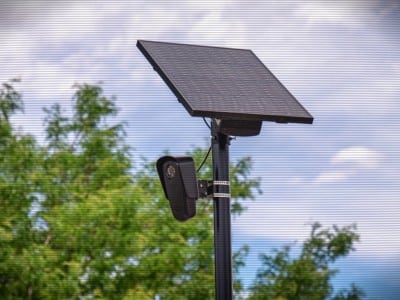 A daylight photo of a Flock automatic license plate reader (ALPR) camera that is centered in the foreground amid a cloudy sky and trees in the background. The image has light CRTV filter lines.