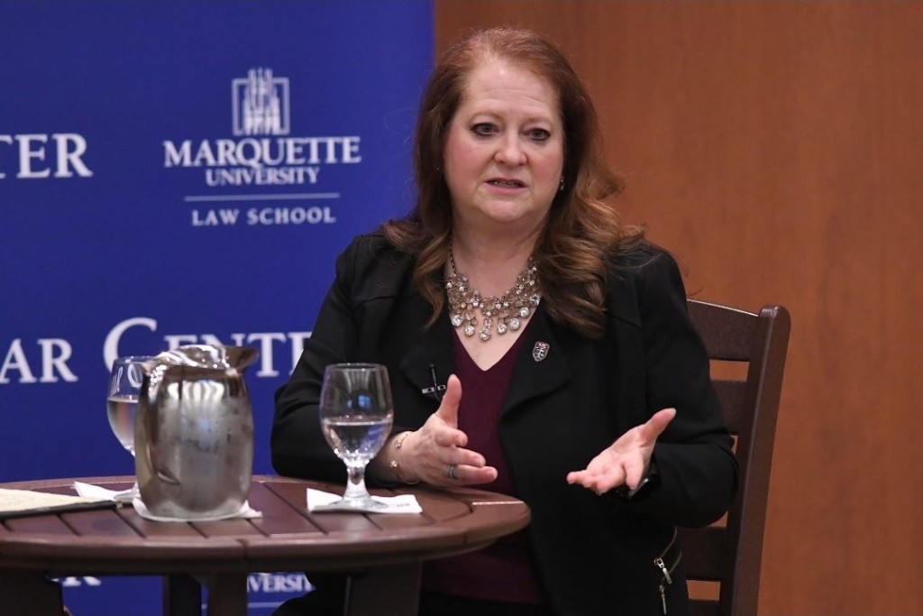 Medium shot of a red-haired woman sitting at a table with a metal pitcher and glass of water. She wears a dark blazer and looks forward, gesturing with her hands. A blue background branded with the Marquette University Law School logo in white appears behind her to the left.
