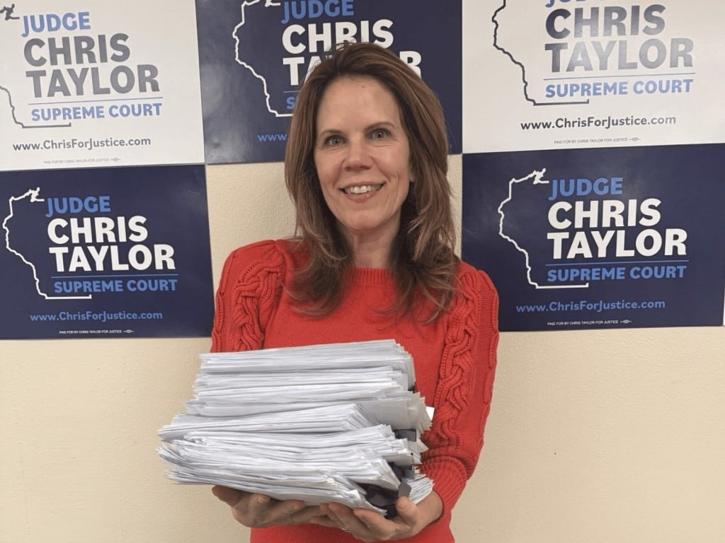 Medium shot of a brunette woman wearing a red sweater and holding a large stack of papers. She smiles and stares directly at the camera. Alternating white and navy blue signs appear in rows on the wall behind her. The signs read "Judge Chris Taylor Supreme Court www.ChrisForJustice.com."