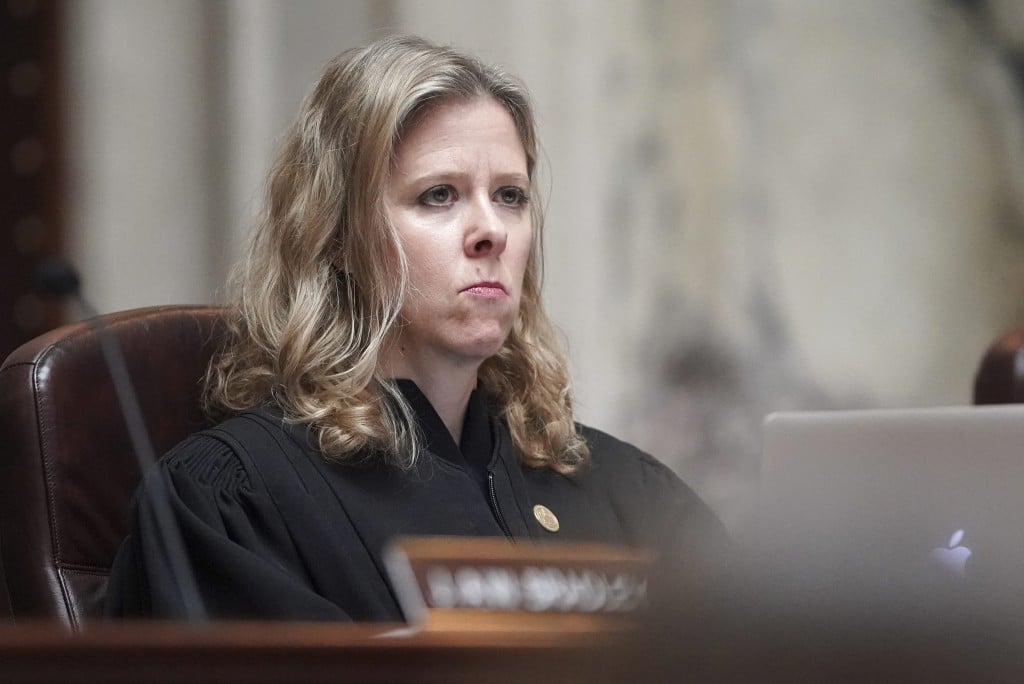 Medium close-up photo of a blonde woman in a black judicial robe sitting at the bench in a courtroom. She stares off to the right with a neutral, observant expression.