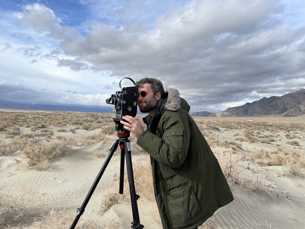 Photo of filmmaker Ben Balcom at a medium shot. He stands in the foreground, wearing sunglasses and an olive-colored winter coat, and looks into the viewing lens of a professional film camera mounted on a tripod. The sandy, expansive area stretches out behind him against a cloudy blue sky and mountains on the horizon.