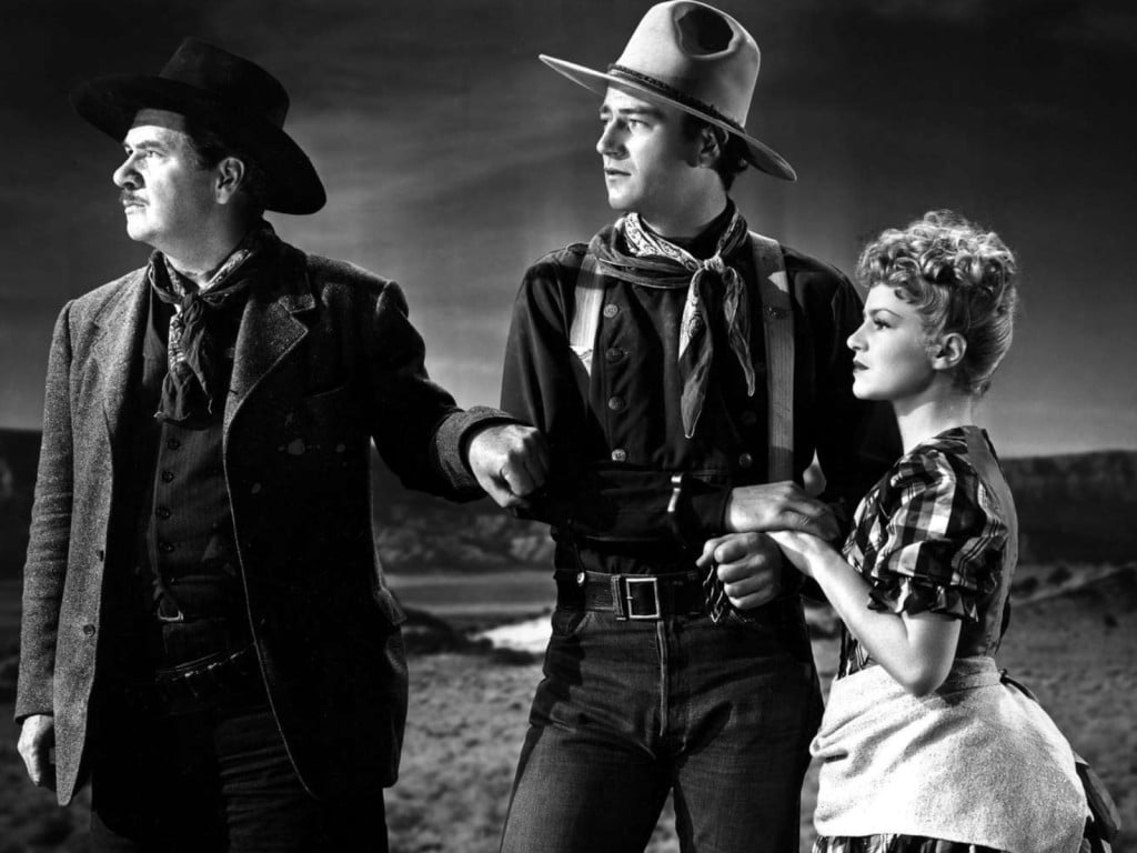 Production photo from the Western "Stagecoach" shows three characters clutching each other's hands and arms. An older man with a moustache and dark cowboy hat stands at the left; a younger, clean-shaven man with a lighter cowboy hat stands taller in the center; and a shorter woman with curly hair stands to the right. They all look off into the distance to the left with neutral expressions.