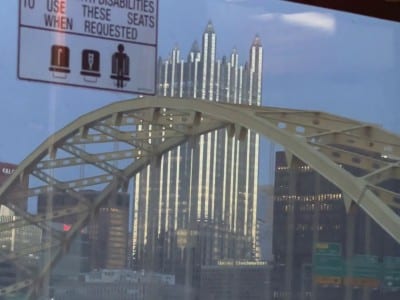 Still from the videopoem "Limerent Pittsburgh" shows a public transit view of a yellow arch of the Roberto Clemente Bridge with skyscrapers and signs of Pittsburgh in the distance, including the massive PPG building, which resembles a cathedral.