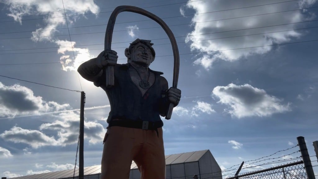 Angled shot of the seven-foot folkloric figure of Joe Magarac against a cloudy blue sky. The statue depicts Magarac with an open workshirt and medallion around his neck, holding a bent steel beam in his hands.