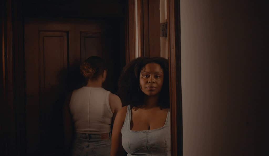 A still from the videopoem "Author's Note" shows a young black woman with curly hair staring directly at the camera while standing in a shadowy doorway. She has a neutral expression. Another woman in a white shirt with her hair pulled back faces away from the camera at the door behind her to the left.