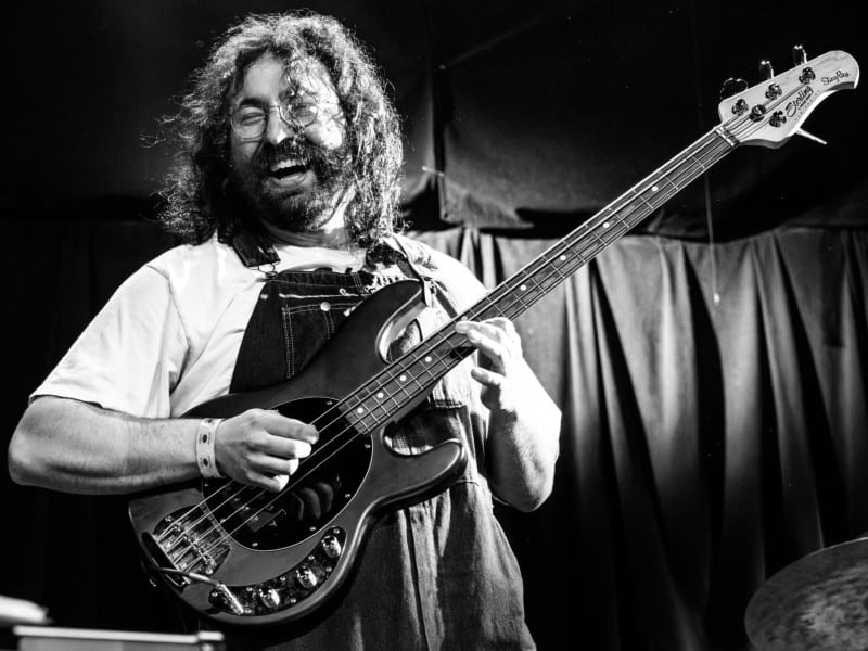Professional black-and-white photo of a long-haired and bearded man smiling widely as he plays an electric bass guitar on a stage. He wears glasses, denim overalls over a white t-shirt, and leans back into the bass groove he's playing.