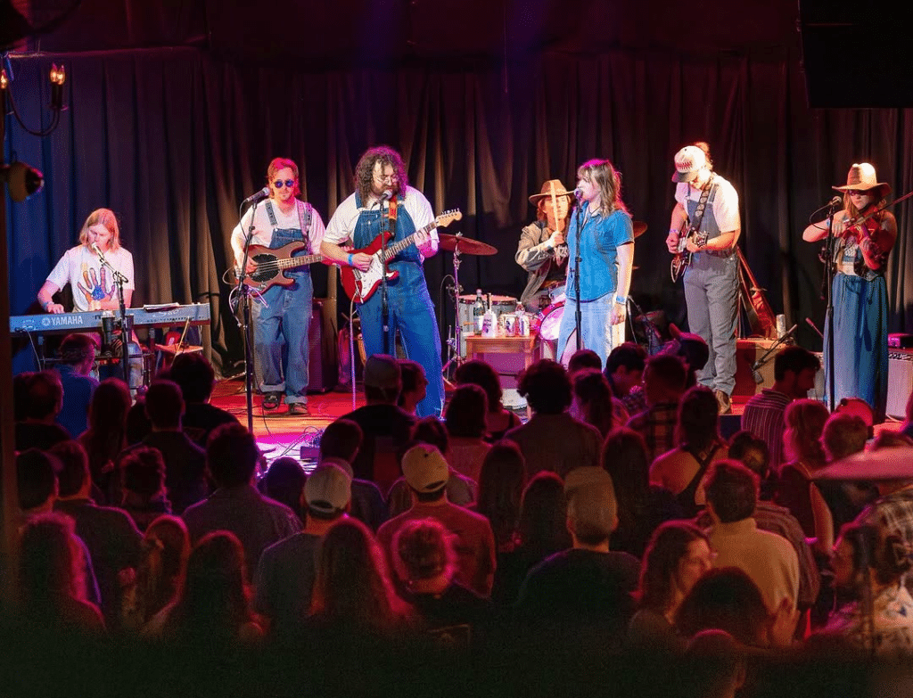 Color photo at a long shot of a septet performing on a stage, with all members facing the camera. The ensemble includes a fiddler, two electric guitarists, an electric bassist, a keyboardist, a drummer, and a vocalist. They play to a large crowd that's packed in near the stage.