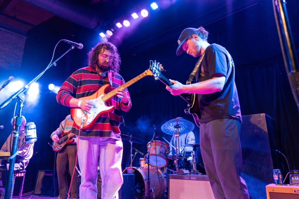 Color photo at a medium shot of a band performing on stage. Two of the male guitars, wearing casual clothes, are centered in the photo in front of the drummer, bassist, and keyboardist partly obscured behind them. Stage lights shine down and concentrate in the left part of the photo.