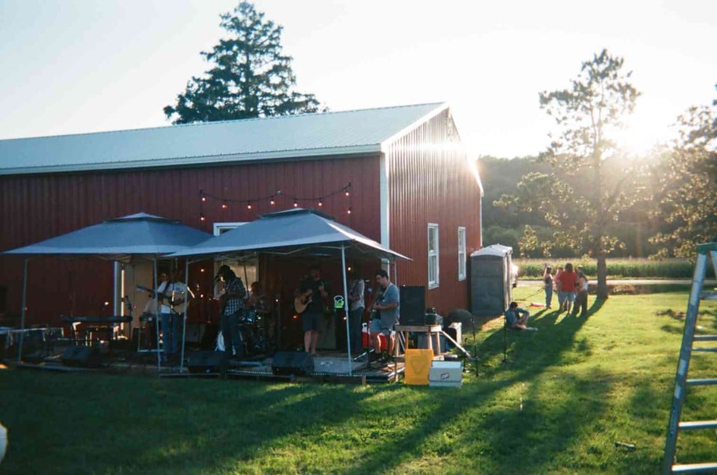 Color photo at a long shot of a sunny, late summer day in a rural area with a large red barn extending from the left side of the frame into the center. People are gathered to the right of the barn and in front of a pond in the background. In the middleground, a band performs directly in front of the barn under two retractable awnings.