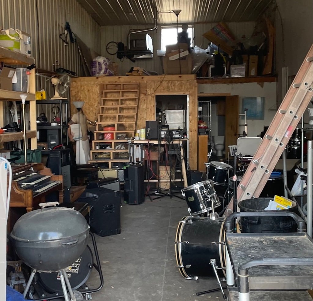 Color photo looking into a storage space that is functioning as a practice space for bands. Several instruments like drums are pushed against the walls but still spill out into the main area. A ladder, a grill, a cart, and other things are also strewn about. In the background, a wood staircase leads up to a second level or loft with other boxes and objects laying about.