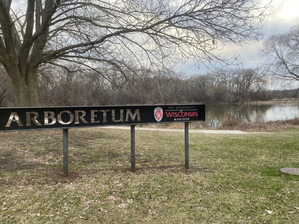 A landscape photo shows the narrow, horizontally elongated entrance sign to the University of Wisconsin–Madison Arboretum in the middleground on a cloudy day in the spring season. The surrounding trees are budding, and a pond extends into the background towards the right of the photo.