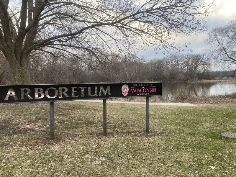 A landscape photo shows the narrow, horizontally elongated entrance sign to the University of Wisconsin-Madison Arboretum in the middleground on a cloudy day in the spring season. The surrounding trees are budding, and a pond extends into the background towards the right of the photo.