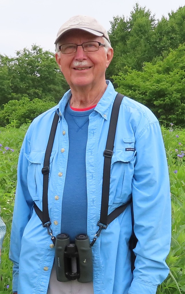 A portrait of an elderly man standing in a lush green field and smiling directly at the camera. He wears an off-white baseball cap, glasses, and an unbuttoned sky-blue dress shirt. He is strapped with a pair of birding binoculars like work suspenders over his clothing.